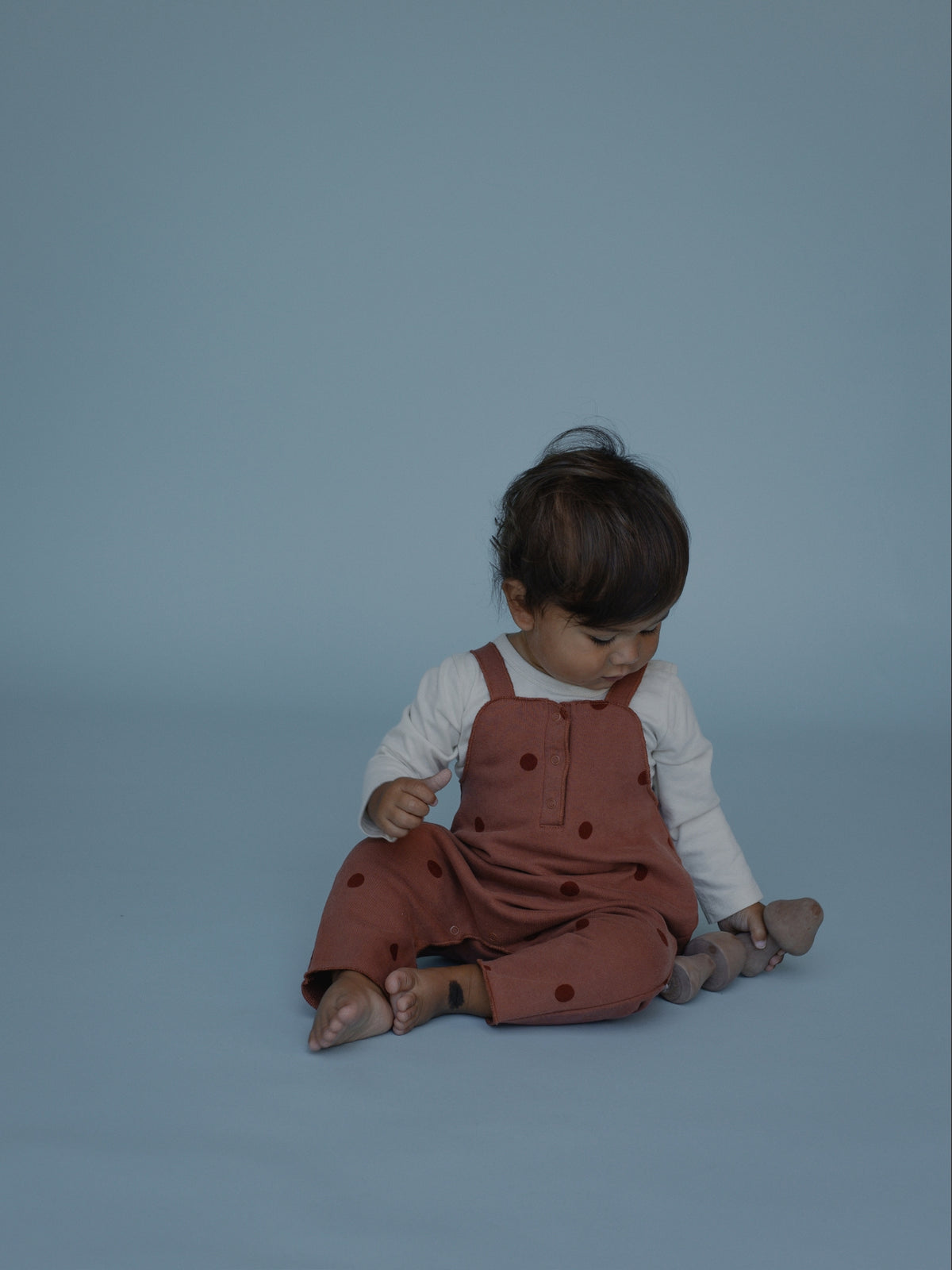 A baby sitting on a solid blue-gray studio background, wearing rust-brown polka-dot overall jumpsuit over a white long-sleeve shirt.