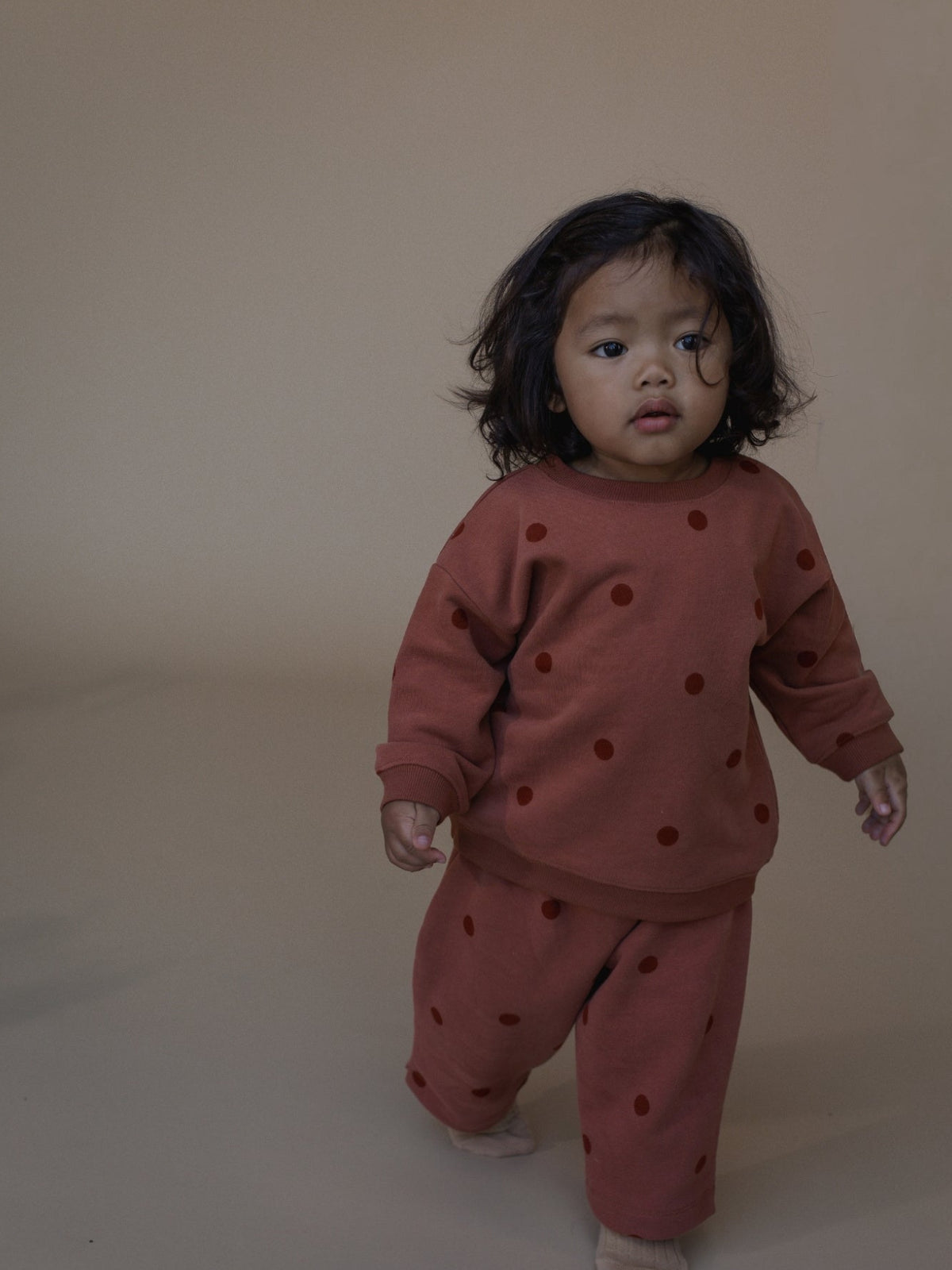 A toddler standing against a plain beige studio backdrop, wearing a matching rust-brown set with dark brown polka dots.