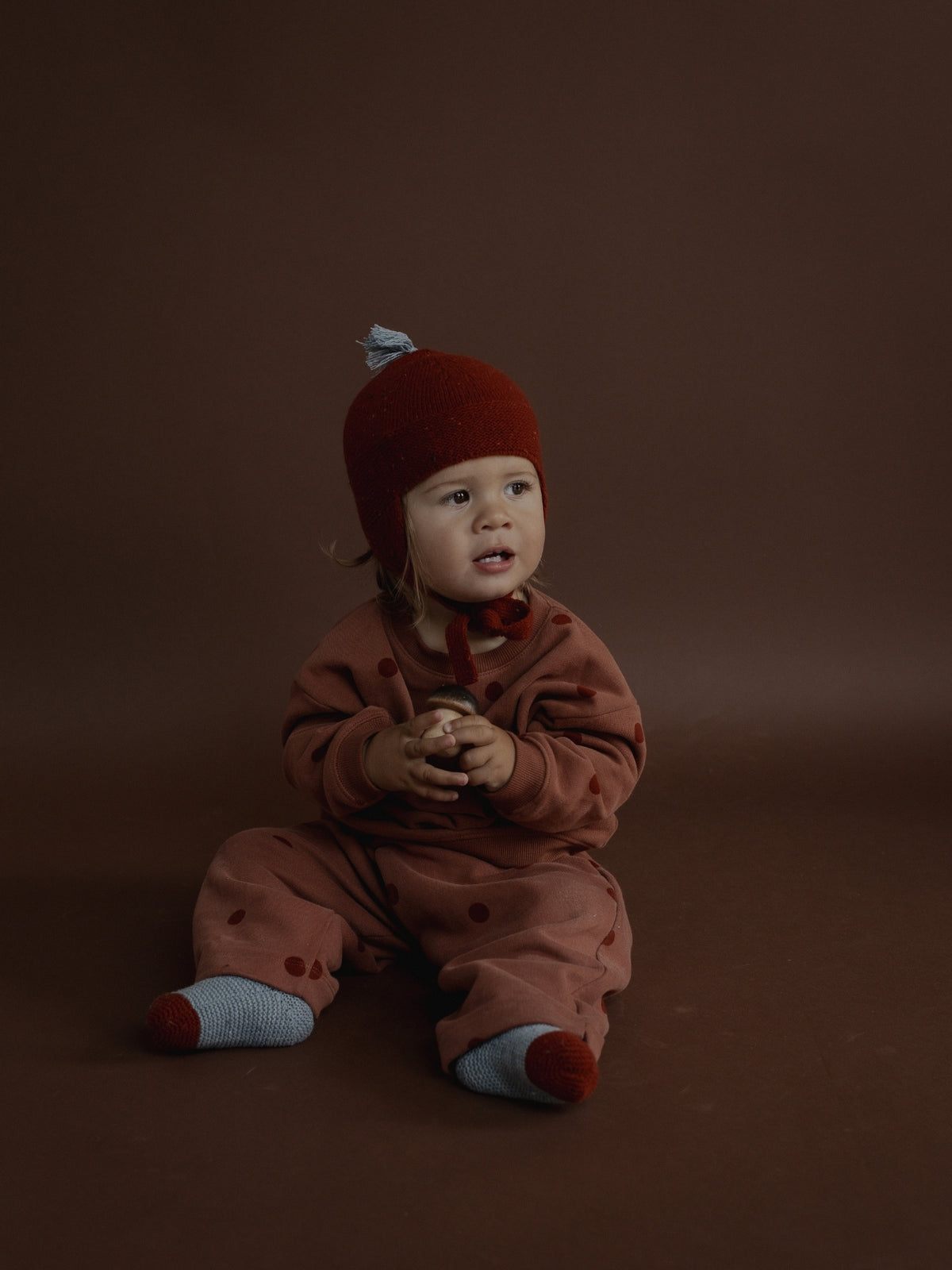 A baby sitting on a dark brown studio background, wearing a matching rust-brown knit hat and footed sleeper with small dark spots, and light blue socks.