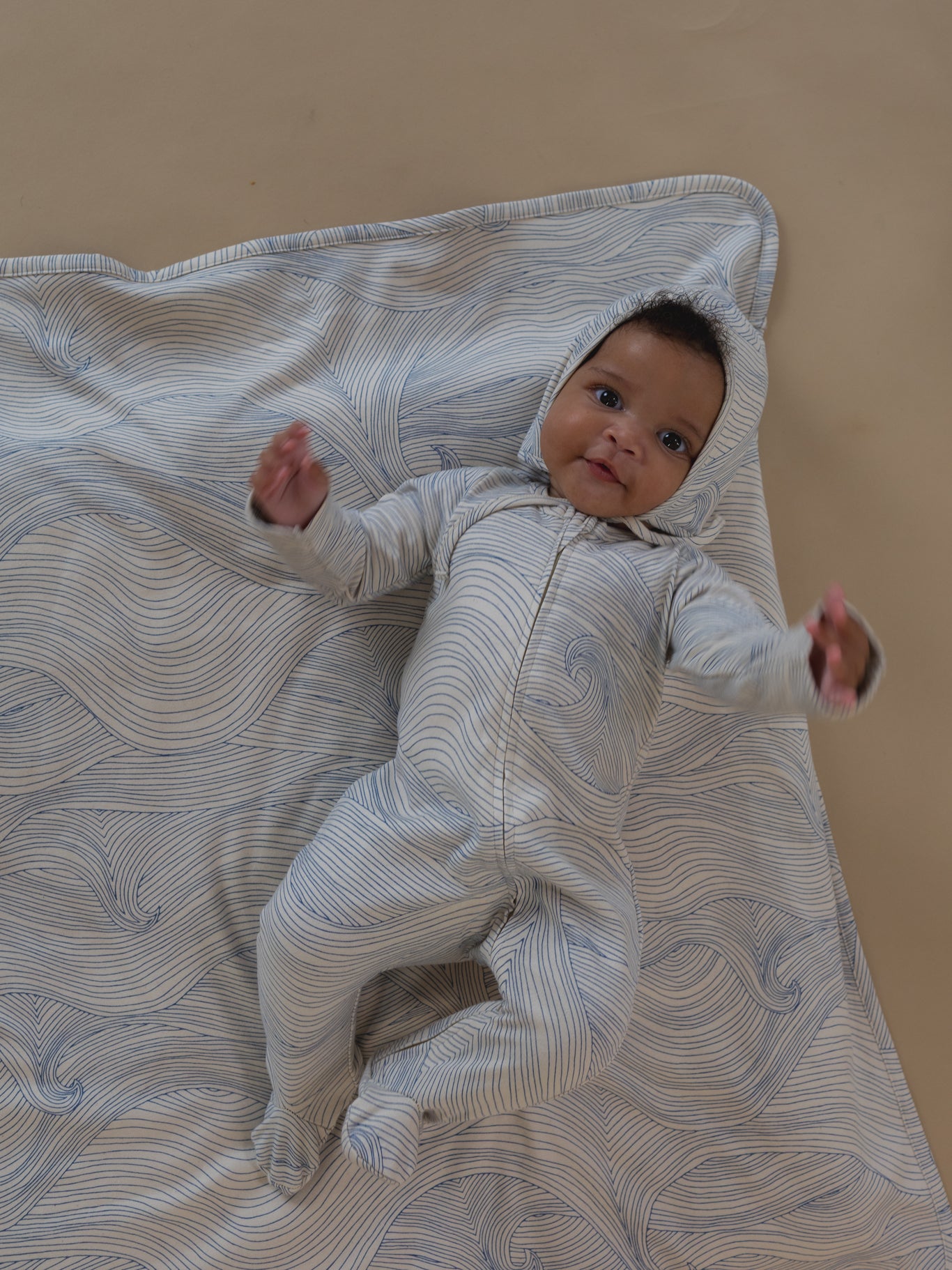 baby in undyed cotton zip sleepsuit with blue line-drawn ocean waves & matching bonnet, lying on blanket