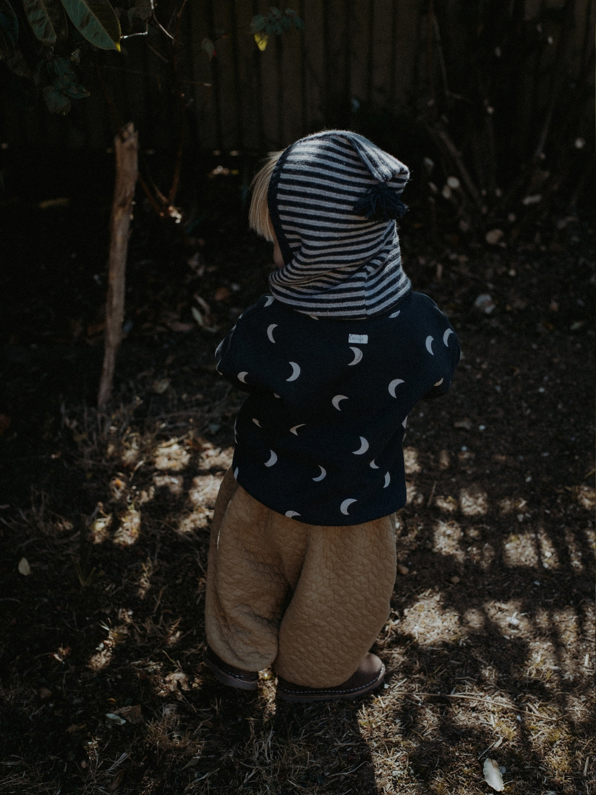 A child standing outdoors in a garden, viewed from behind, wearing a navy long-sleeve top with a white crescent moon pattern, tan pants, and a striped knit bonnet.