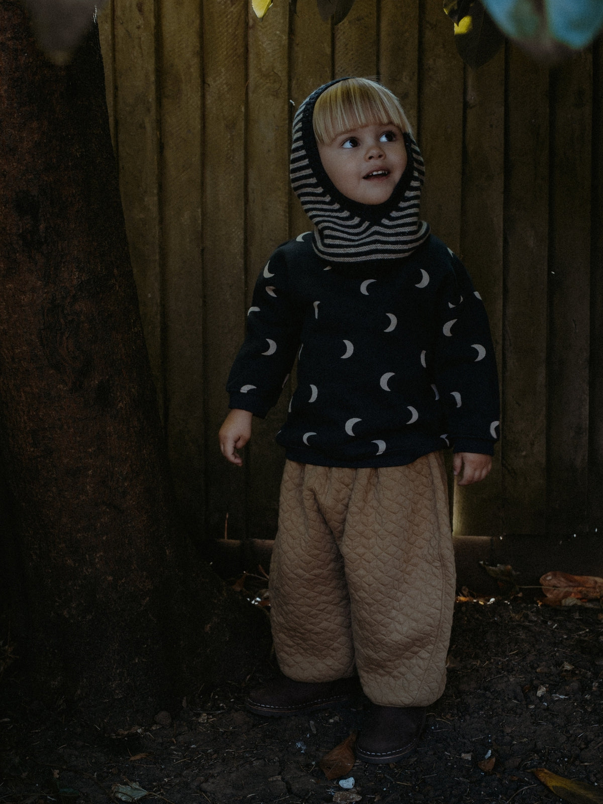 A young child wearing a dark navy fleece hoodie with a white crescent moon pattern, a striped hood, and quilted tan pants, standing outdoors against a wooden fence.
