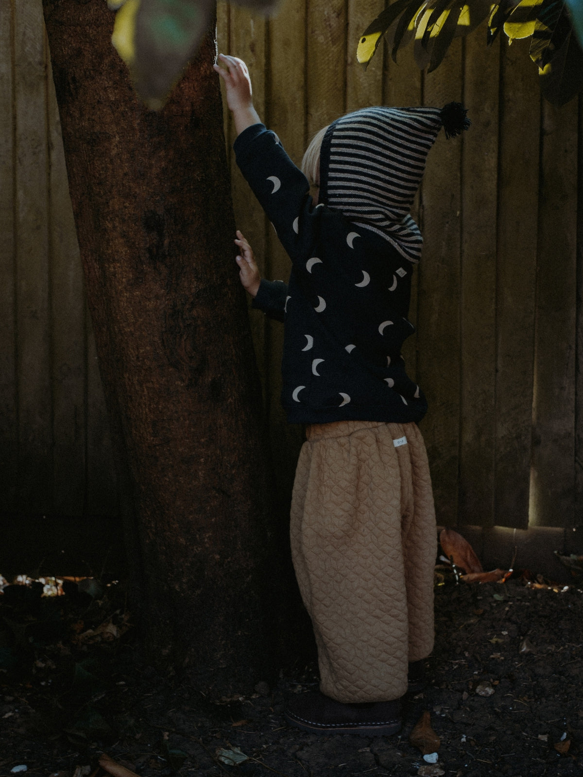 A child wearing a dark navy long-sleeve top with a white crescent moon pattern, beige quilted pants, and a striped beanie stands next to a large tree in front of a wooden fence.