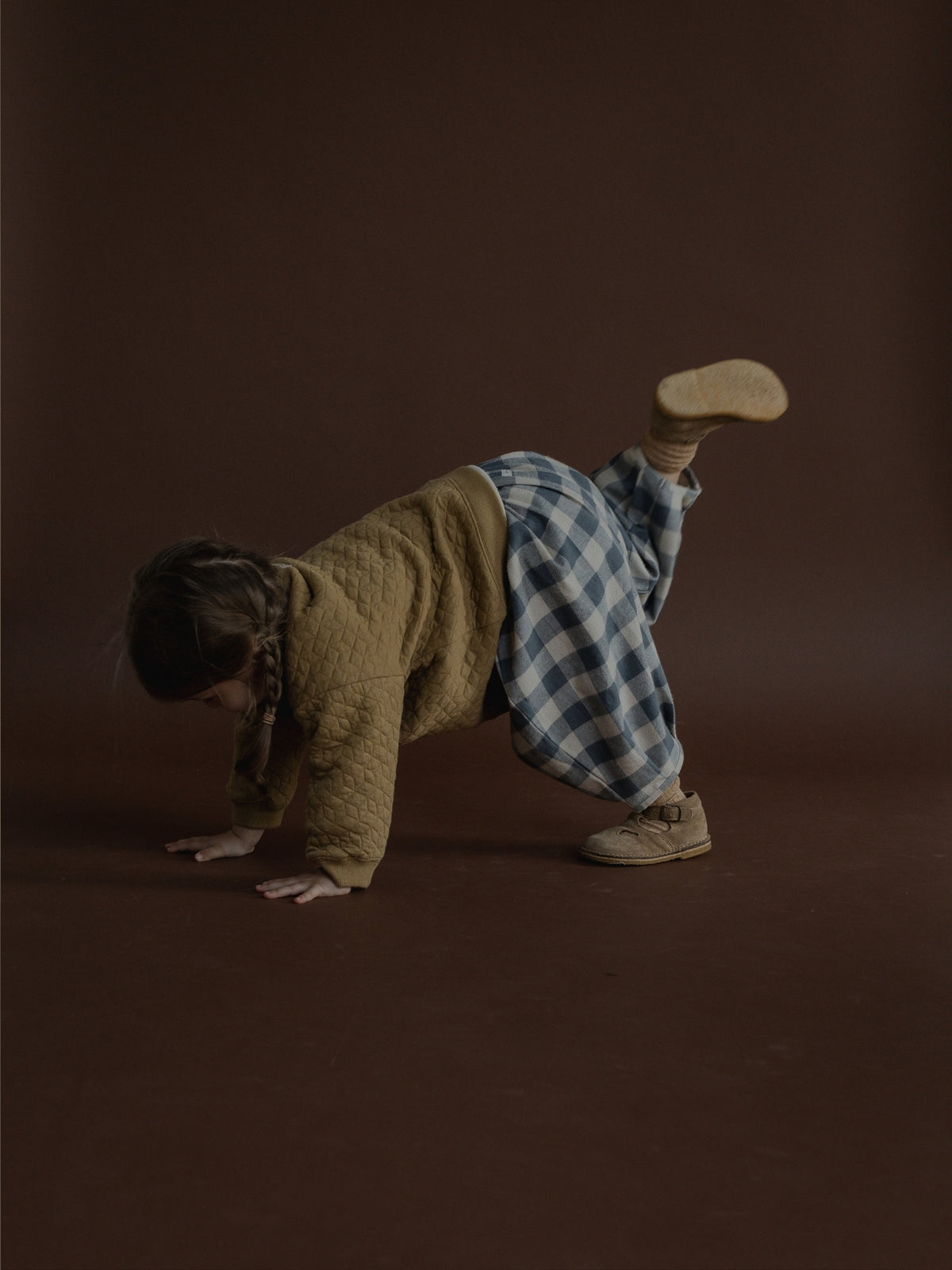 A child wearing a textured olive green top, a blue and white checkered skirt, and beige shoes poses on hands and knees against a solid dark brown studio background.