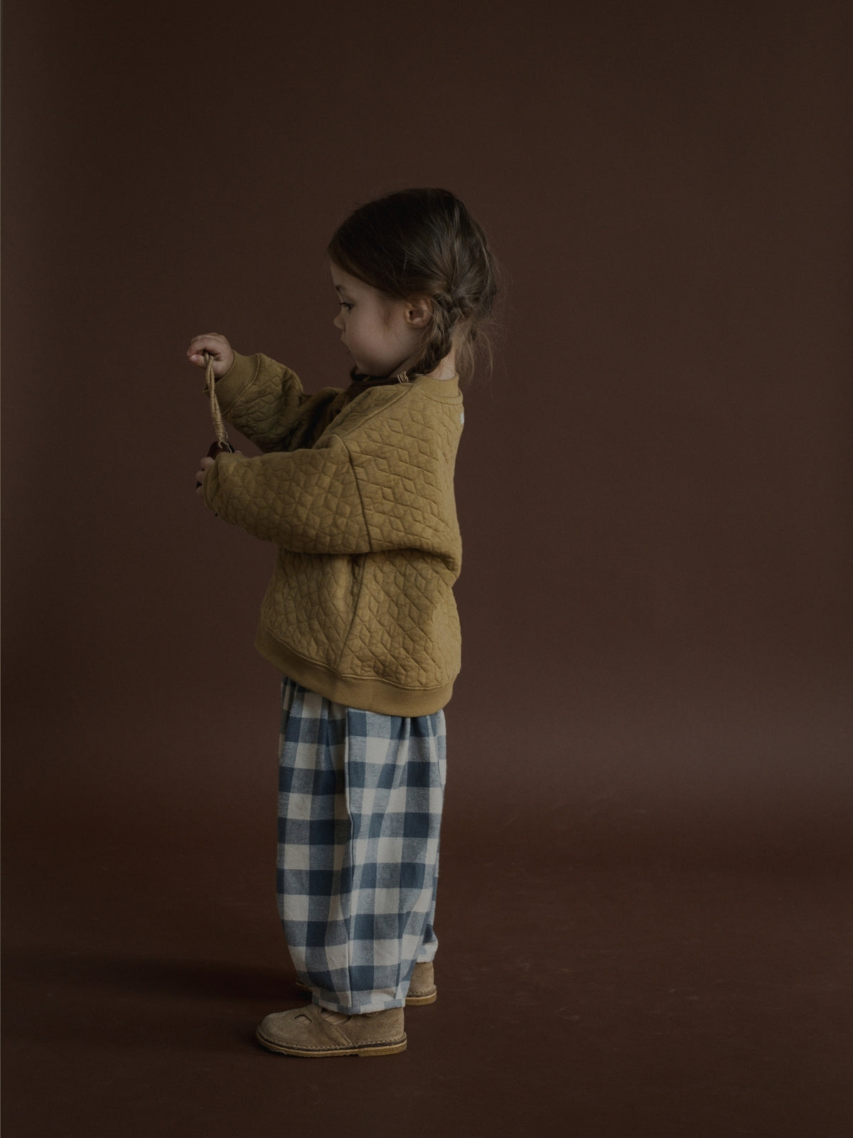A young child stands in profile against a dark brown studio backdrop, wearing a mustard yellow quilted knit sweater and blue-and-white gingham pants.