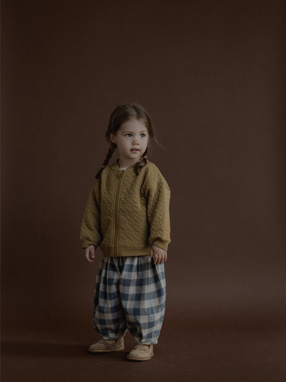 A young child stands against a solid dark brown studio backdrop, wearing a textured mustard-brown zip-up jacket and loose blue-and-white plaid pants.