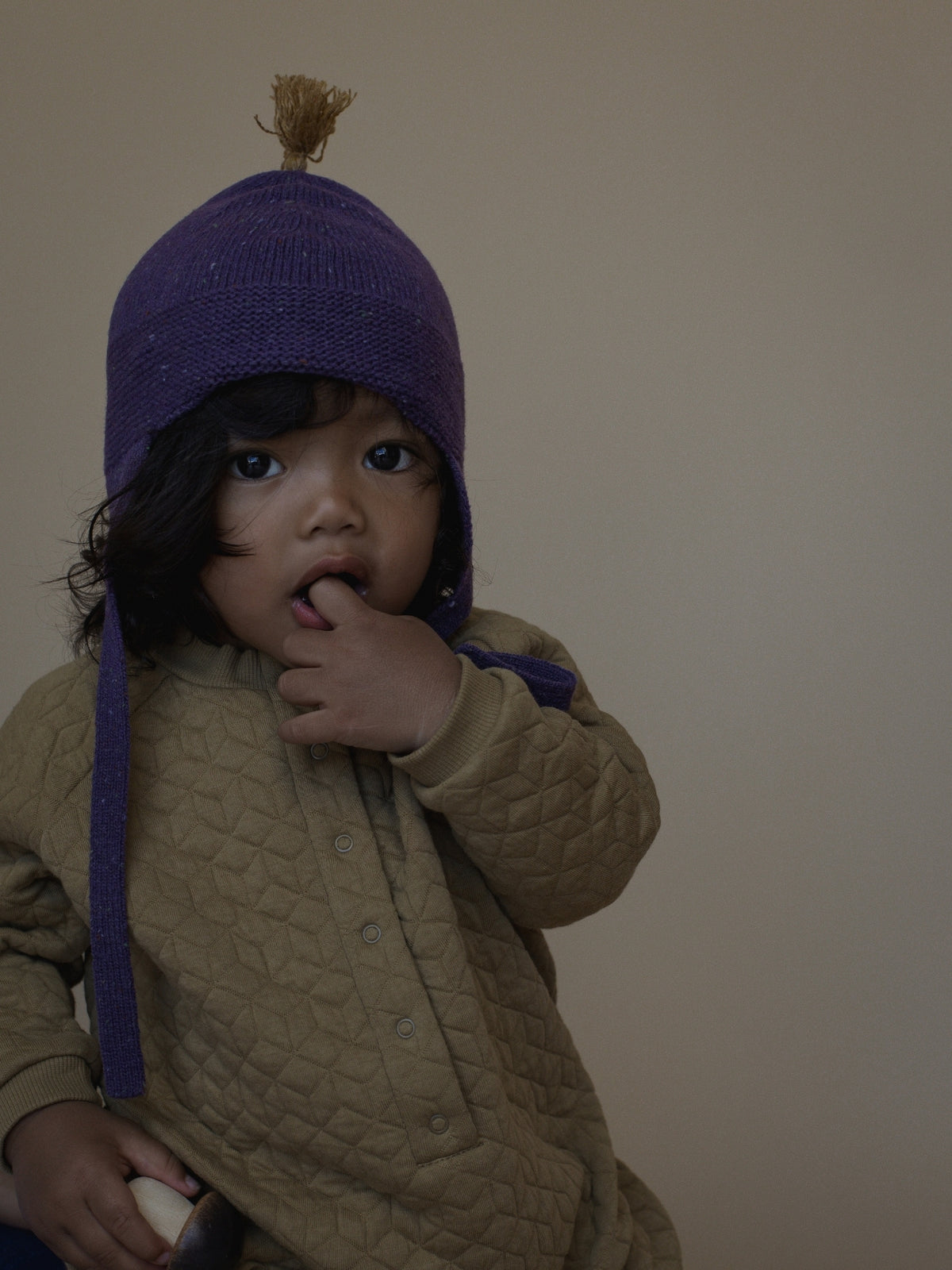 A child wearing a purple knit bonnet with a small tan tassel and a quilted tan jacket, posed against a plain beige wall.