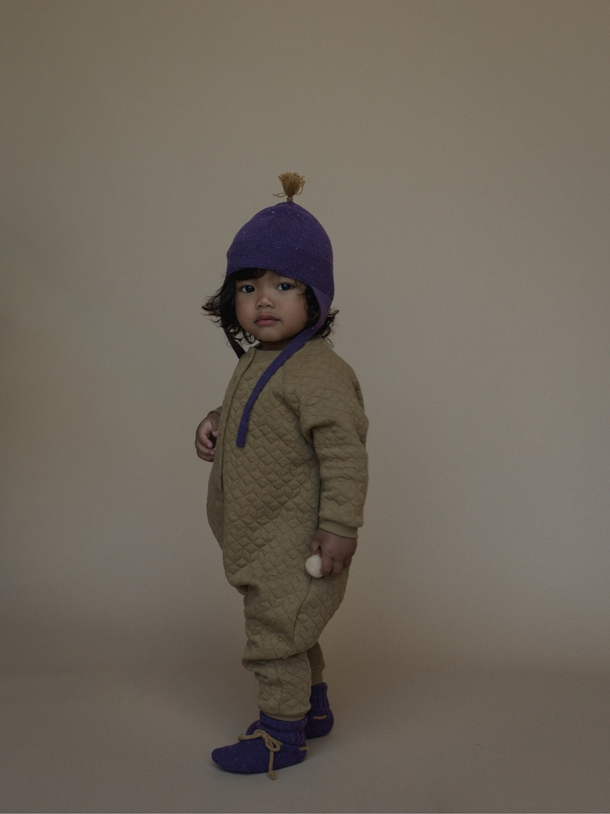 A toddler stands in profile wearing a quilted one-piece jumpsuit in taupe-beige with purple trim, a purple knit hat with a tan pom-pom, and purple booties, against a plain warm beige studio background.
