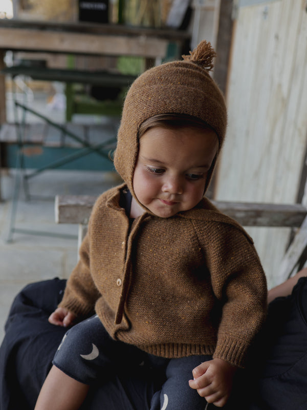 smiling baby wearing matching knitwear brown wool cardigan & hat with tassel sitting 