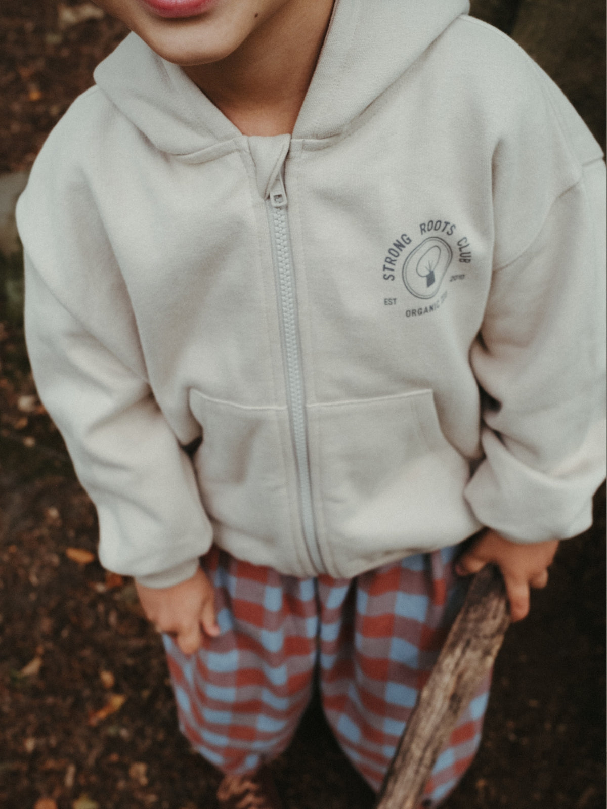 A child wearing a cream-colored zip-up hoodie with a circular 'Strong Roots Farm' logo on the left chest, paired with red and blue checkered pants, standing outdoors on a dirt ground.