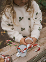 close up of toddler in organic cotton christmas holly & candy cane cream sweatshirt holding cup of cocoa