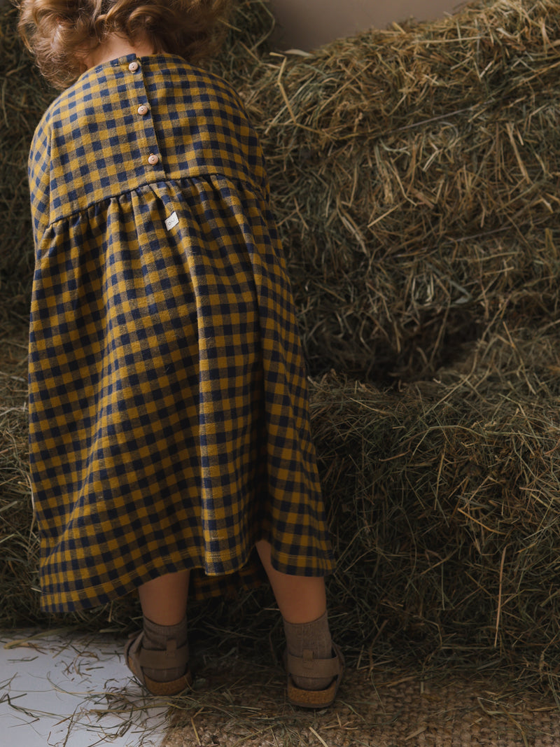 toddler in gingham dress with buttons on the back & gathered waist & brown sandals handling straw crops