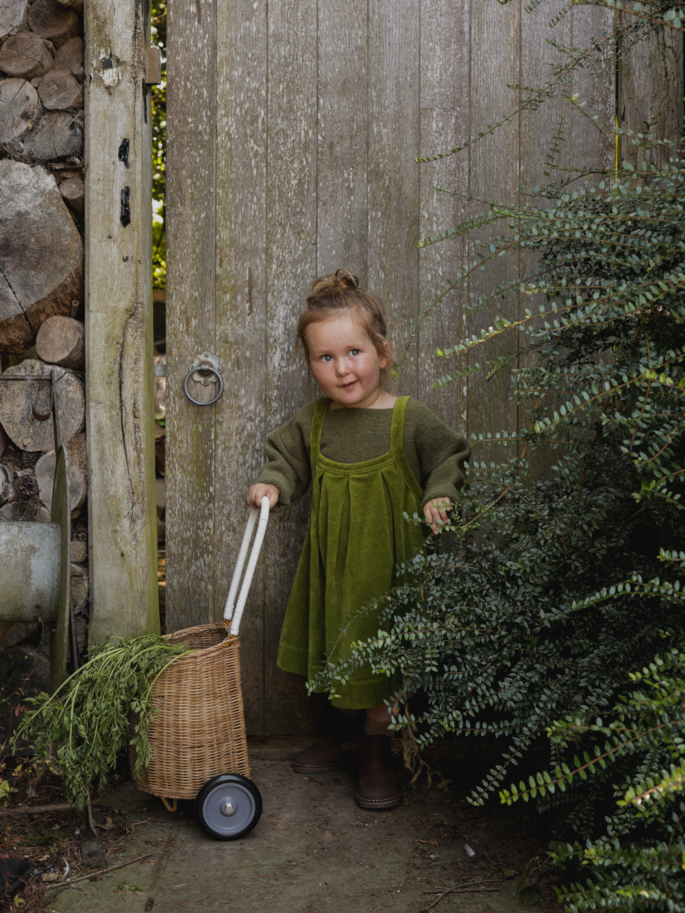 toddler in pinafore skirt in pea pod green & sweatshirt pushing rattan trolley with carrots