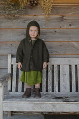 Toddler Wearing Boxy Wool Hooded Jacket In Basil Green Standing On Garden Bench