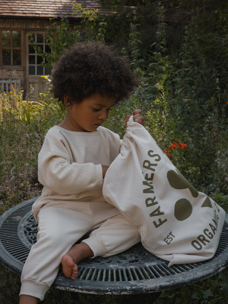 toddler wearing cream sweatshirt and sweatpants sitting on patio table searching drawstring bag