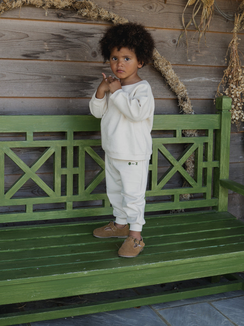 toddler in matching undyed cotton cream sweatshirt sweatpants & brown shoes standing on green bench