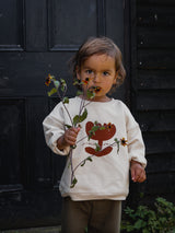 toddler dressed in cream sweatshirt with plant & slogan in red & waffle long pants holding flowers