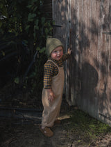 toddler in navy & yellow gingham shirt & beige oversized dungarees green beanie with tassel in a garden