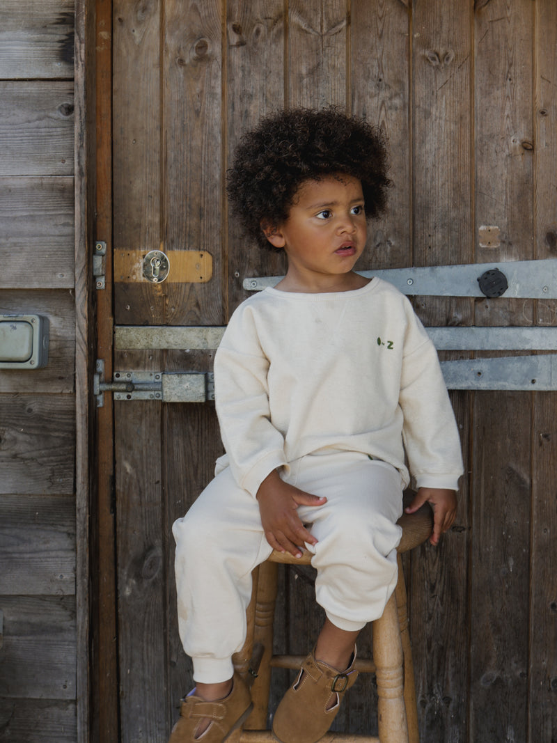 toddler in matching undyed cotton cream sweatshirt & sweatpants with embroidered text sitting on stool