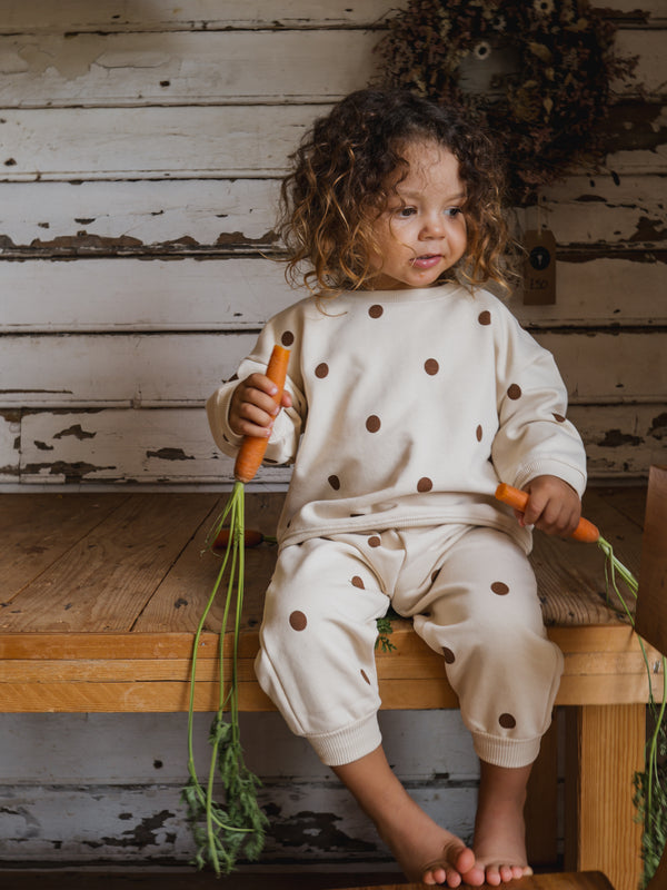 toddler in matching cream sweatpants & sweatshirt with brown dots sitting on wooden table holding carrots