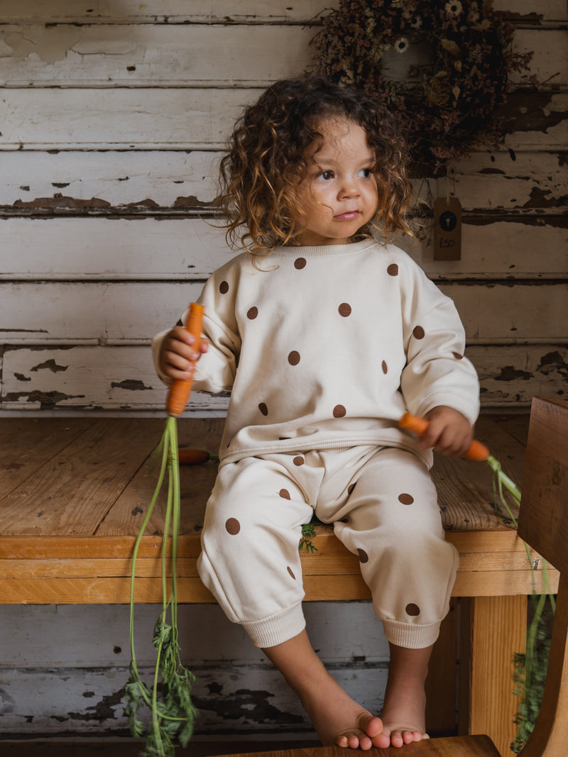 toddler in cream sweatshirt & sweatpants set with brown dots sitting on wooden table holding carrots