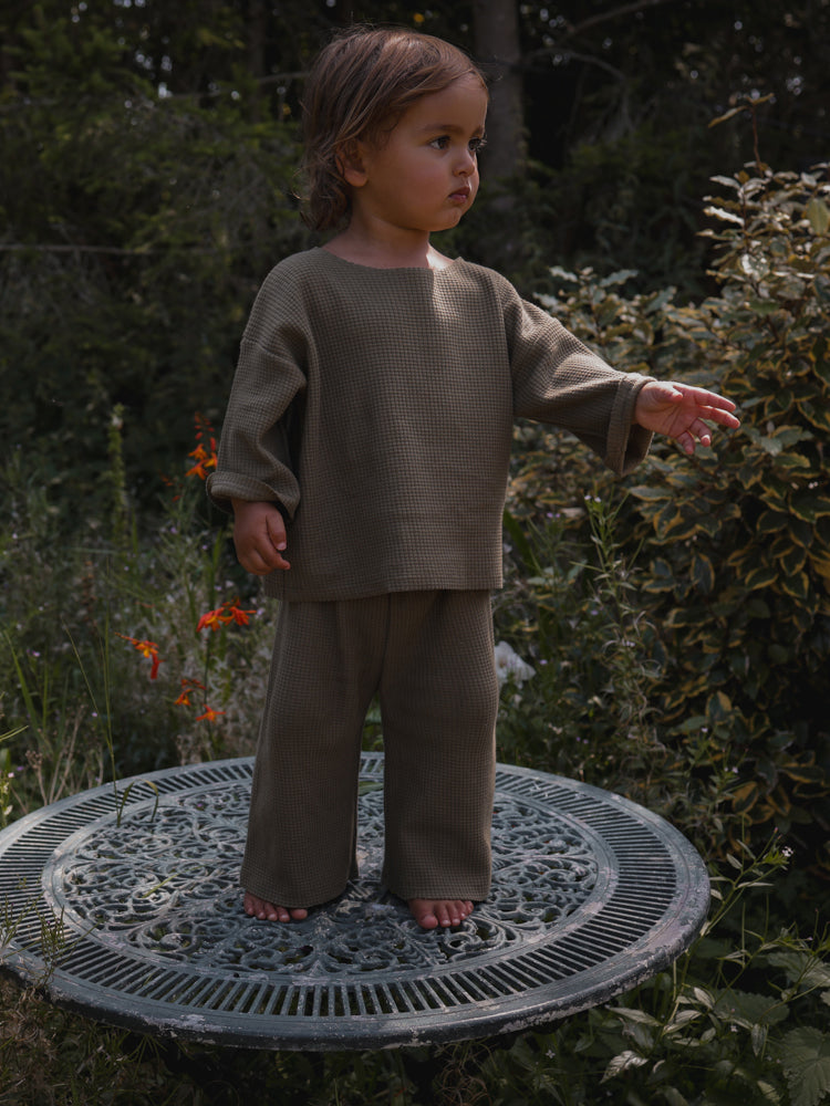 toddler dressed in matching olive sweatshirt & wide leg pants standing barefoot on garden table