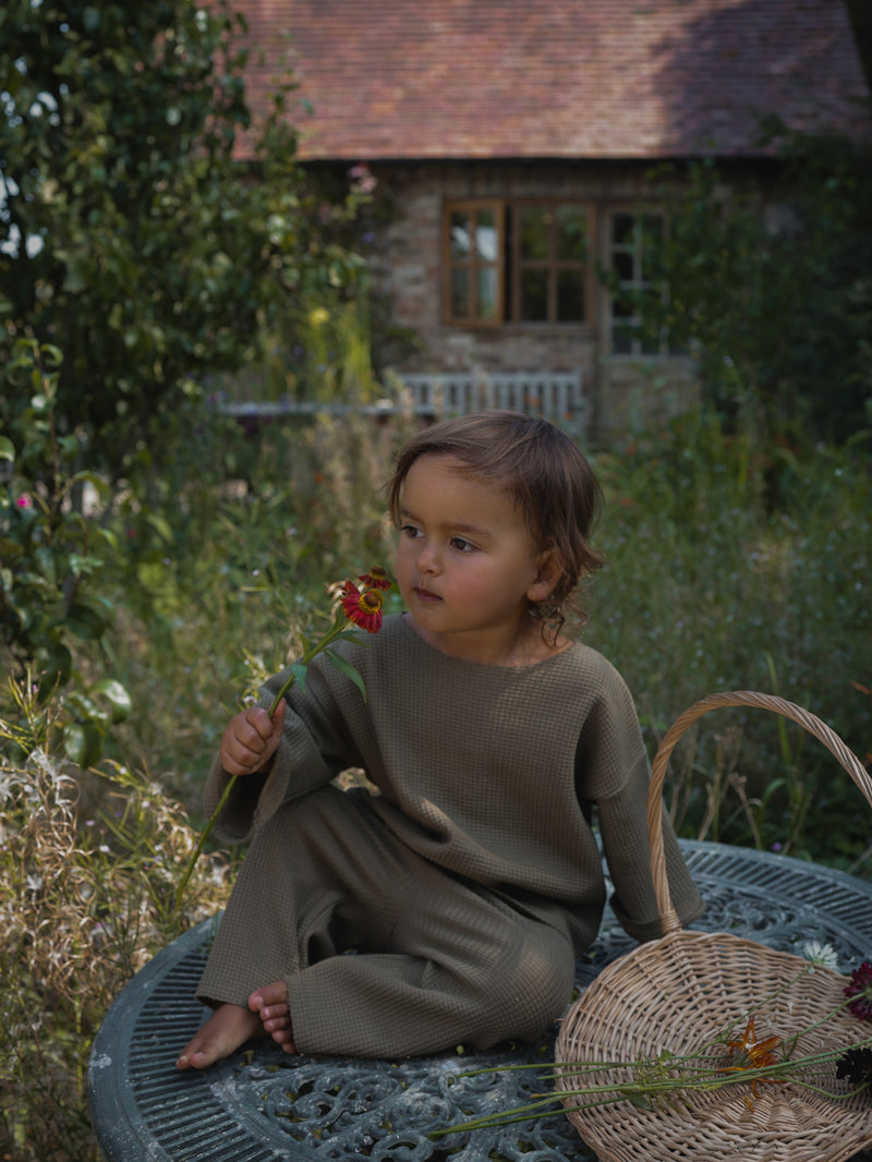 toddler in olive wide leg pants & boxy sweatshirt sitting on table with wicker basket holding flower stalk