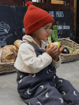 toddler in paprika red beanie with tassel cream fleece sweatshirt & oversized black dungarees eating pear