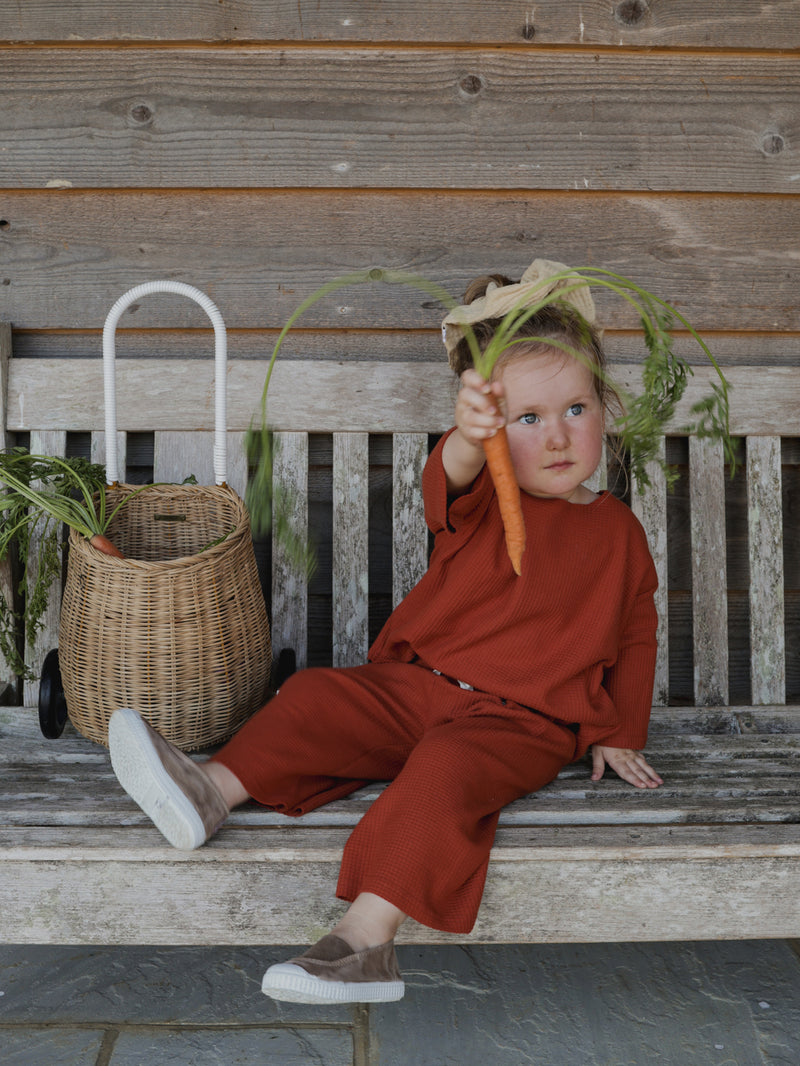 toddler in paprika red cotton waffle boxy sweatshirt & pants sitting beside wicker trolley holding carrot