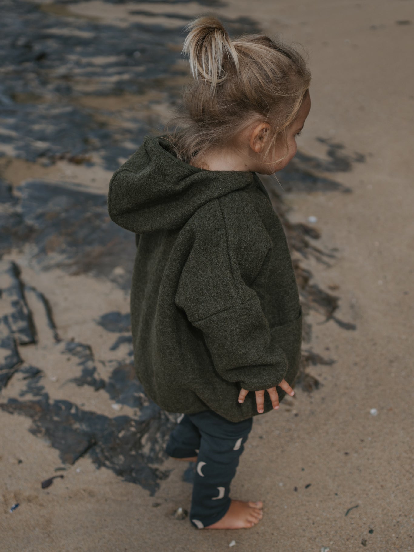 Child wearing a green wool hoodie standing on a sandy beach.