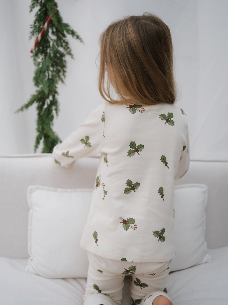 back view of toddler in matching organic jersey cream pyjamas with holly & candy cane kneeling on sofa