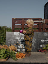 toddler in matching navy & yellow gingham shirt & pants set standing in front of fresh produce holding aubergine