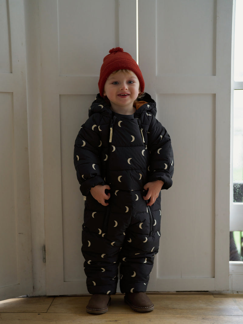 toddler in winter midnight padded snowsuit with white moons & hood with red wool beanie standing at door