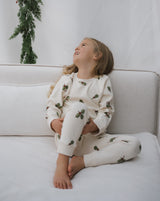 toddler wearing organic jersey pyjamas in cream with holly & candy cane sitting with leg up on sofa