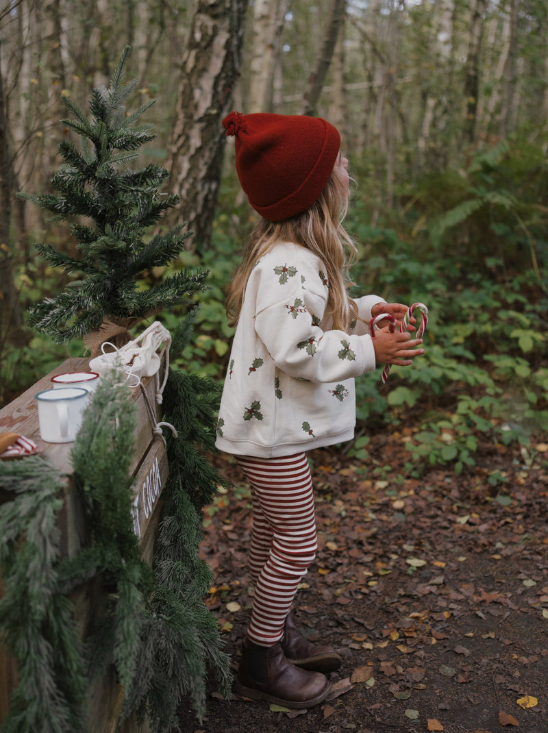 toddler in cream sweatshirt with holly & candy cane stripes leggings beanie boots in front of cocoa stand