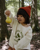 toddler in paprika red beanie & cream logo sweatshirt holding a pear in a forest