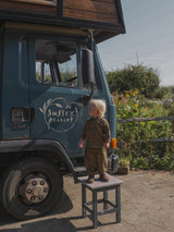 toddler in navy & yellow flannel shirt & pants set standing on stool in front of farm produce truck