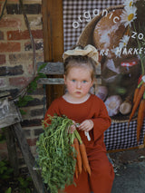 toddler in red long-sleeve shirt & pants with hair bundled up in scrunchie holding bunch of carrots