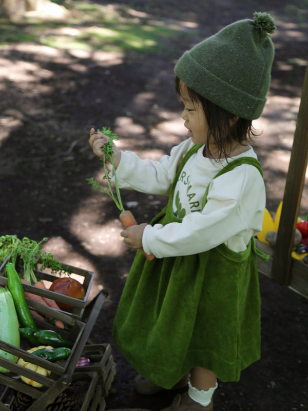 toddler in green wool beanie cream sweatshirt & light green skirt holding carrot