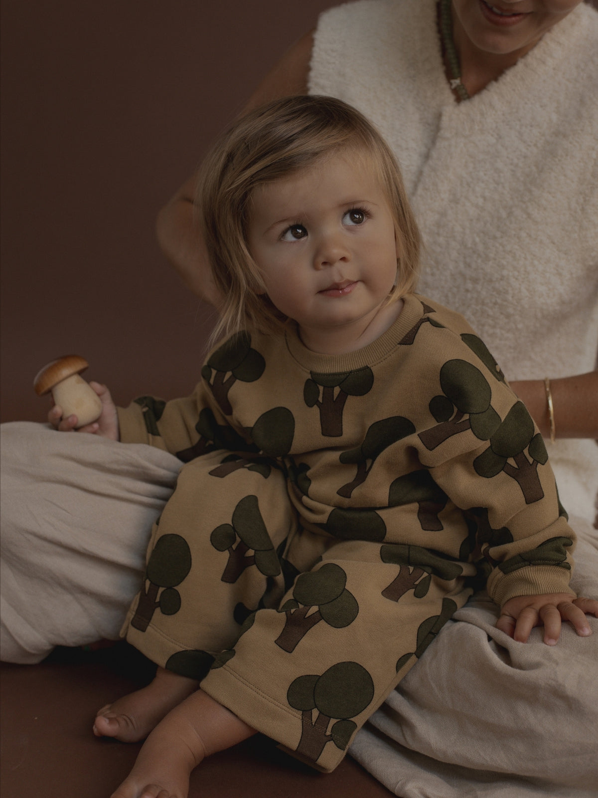 A toddler wearing a tan and olive green tree-print set sits on a soft cream-colored blanket, with a woman in a fuzzy cream sweater sitting behind them against a dark brown background.