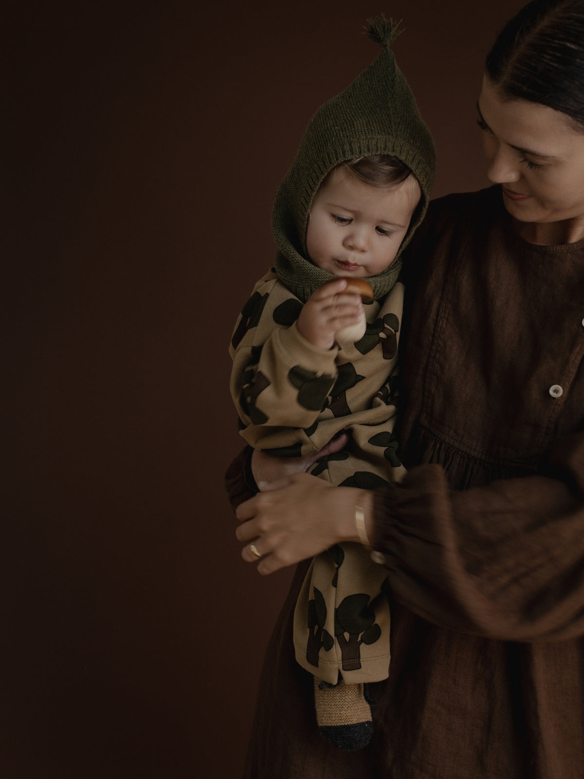 A baby wearing a brown and beige tree-patterned outfit and an olive green knit bonnet, held by an adult in a matching brown dress against a dark brown background.