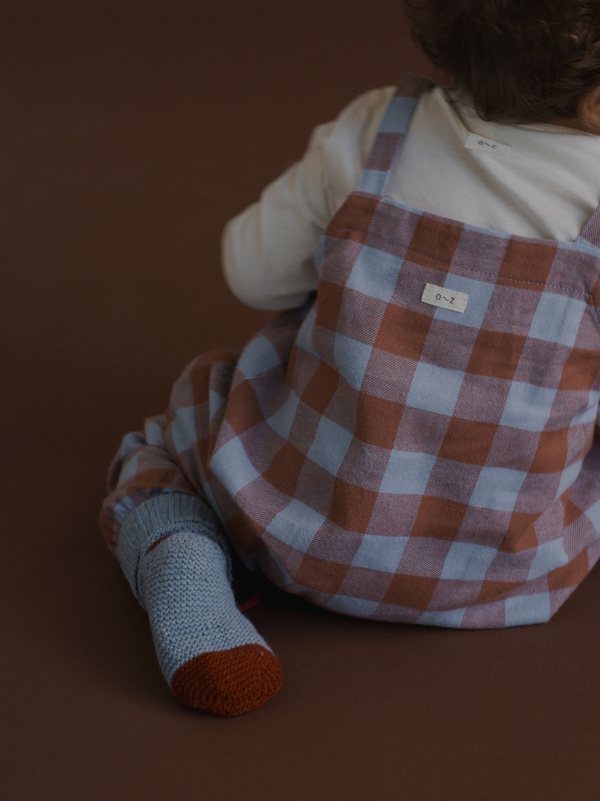 A child wearing a brown and light blue gingham check overalls outfit, sitting on a solid brown surface. The child is also wearing a white long-sleeve shirt and a light blue knit sock with a brown toe.