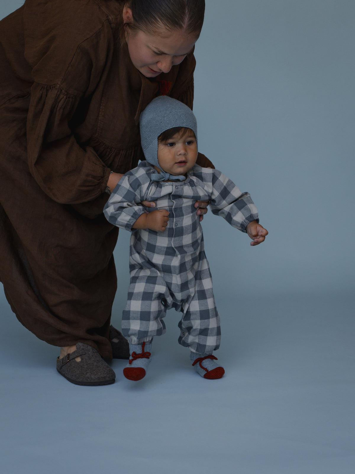 A baby wearing a gray and white checkered one-piece jumpsuit with a matching knit bonnet and red shoes, being supported by an adult in a brown dress against a solid blue-gray studio background.