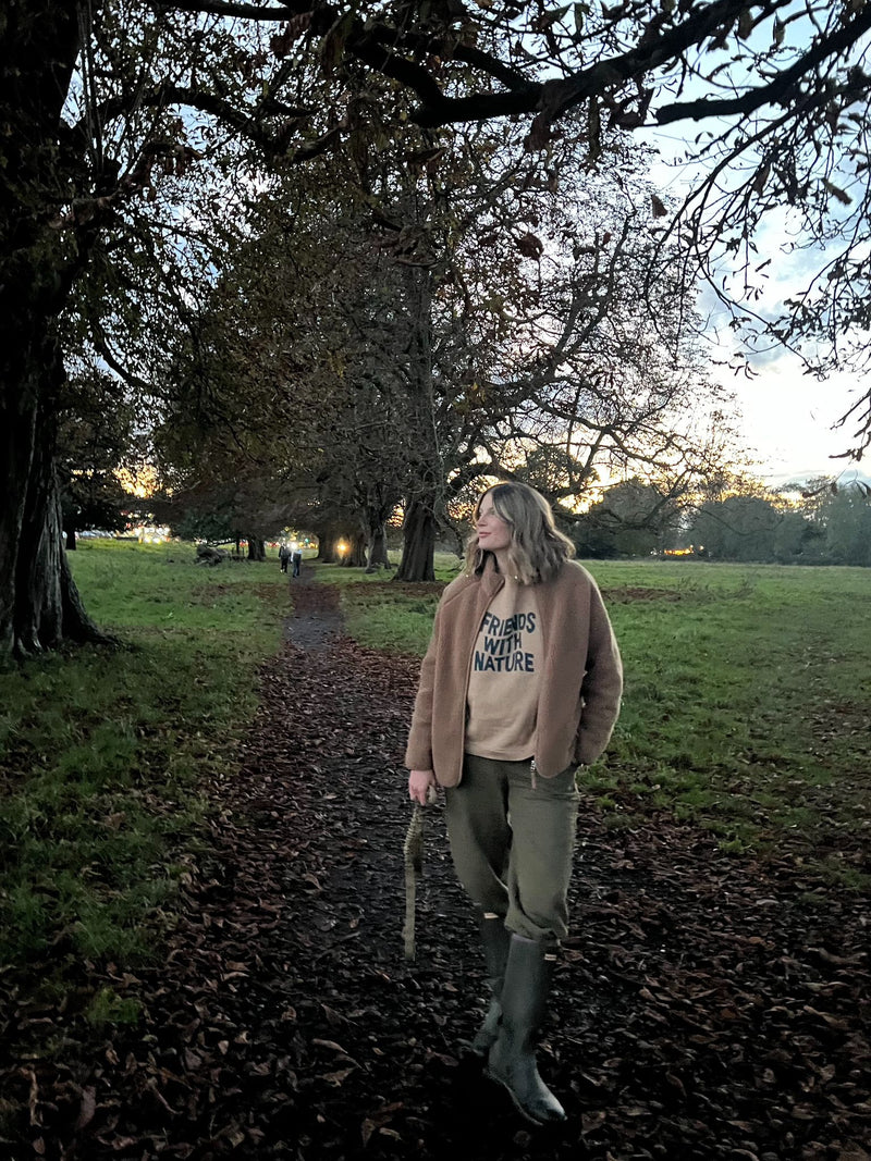 woman in brown sweatshirt with FRIENDS WITH NATURE text & jacket pants boots standing in park