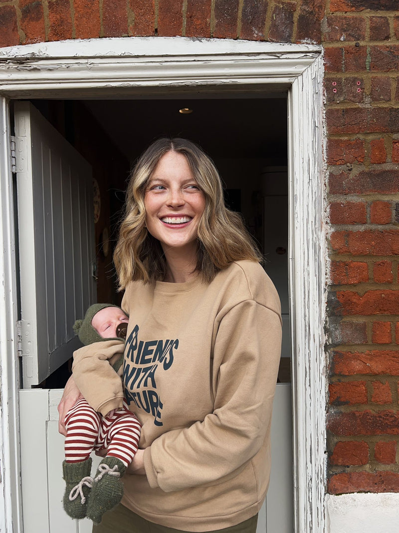 woman in brown sweatshirt with text carrying sleeping baby in matching sweatshirt leggings wool beanie & booties