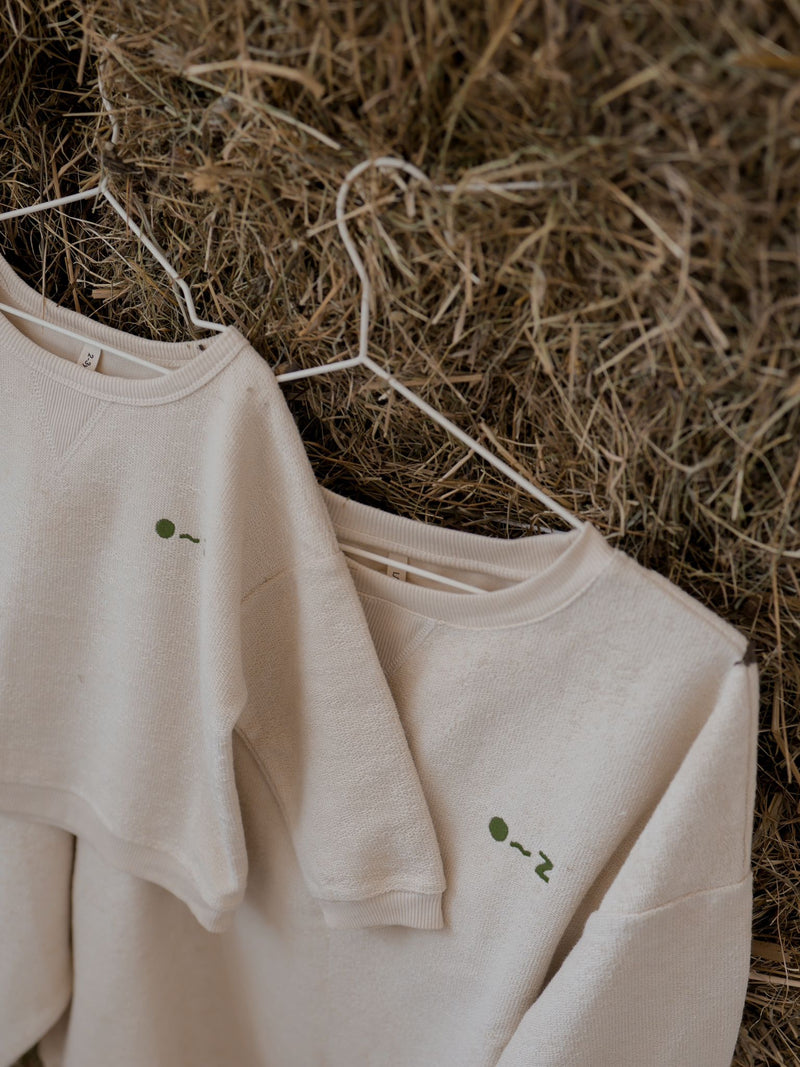 close up of women's & toddler cream sweatshirt with embroidered O~Z text hanging on straw bale wall