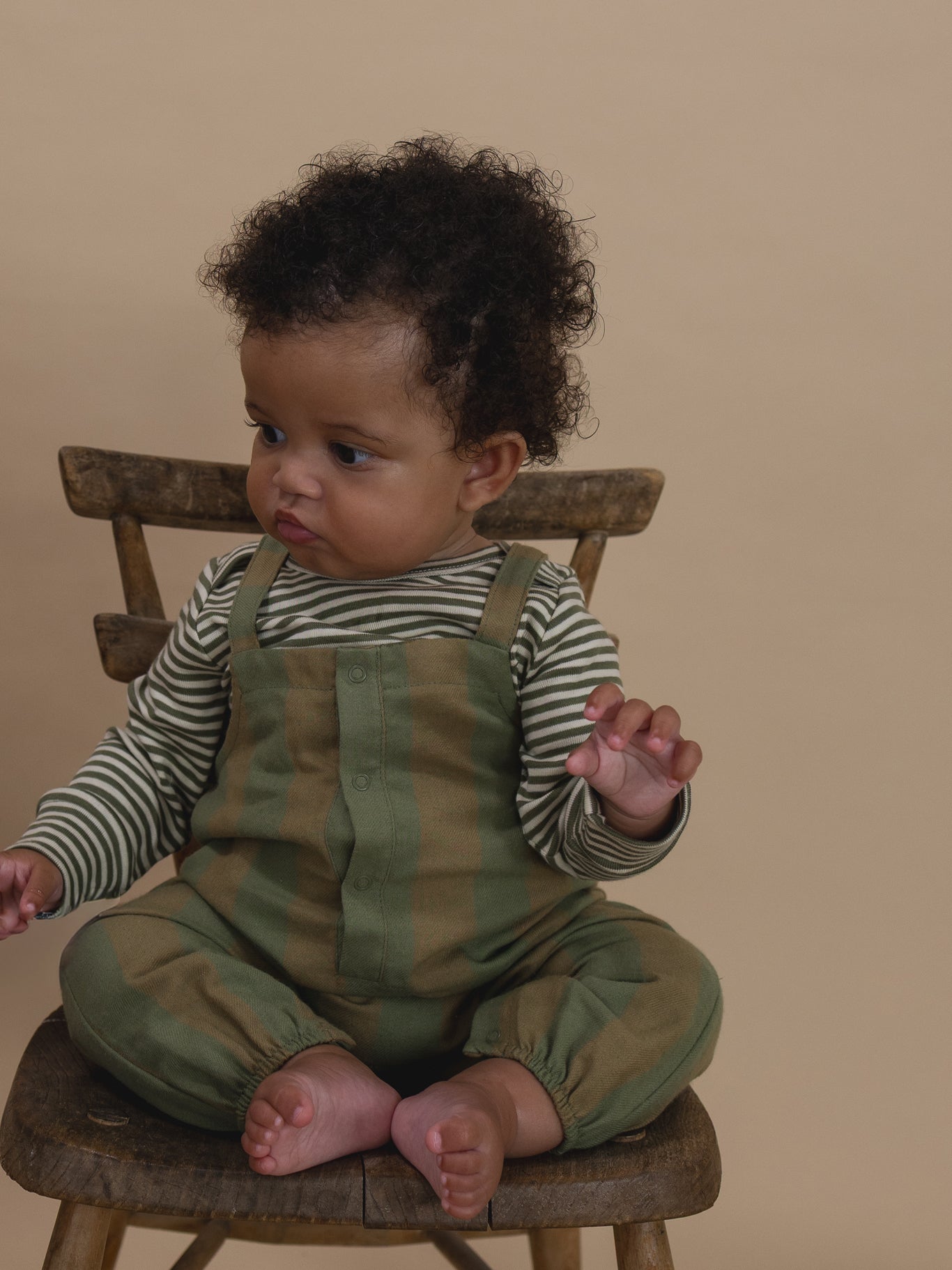 baby in green and cream gingham salopette with gathered cuffs, and striped sweatshirt sitting on chair